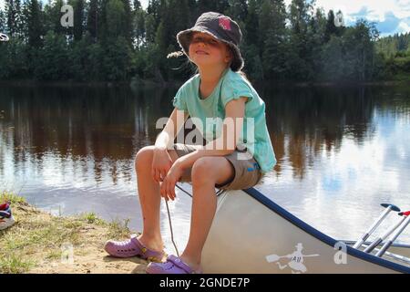 a girl sitting on a canoe laying on the river bank of the Klarälven river in Sweden,  the girl is chewing on a straw of grass Stock Photo