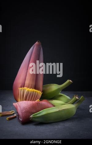 ash plantains or green raw banana with banana flower also known as banana blossom, vegetables isolated on a dark background, closeup view Stock Photo