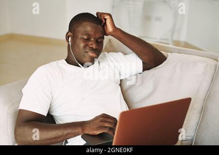 a man of african appearance with a closed laptop sits on the couch and listens to music on headphones Stock Photo