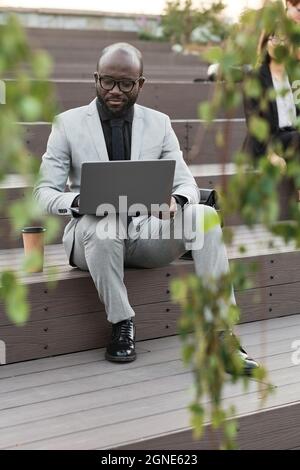 Vertical full body shot of young adult African American man wearing stylish suit sitting outdoors with laptop working on business plan Stock Photo