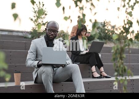 Horizontal shot of modern man and women sitting outdoors on stair benches working on laptops during coffee break time Stock Photo