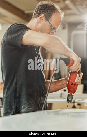 Carpenter drilling a hole in a large panel of wood using a hand held red power drill in a woodworking factory or workshop during the production proces Stock Photo