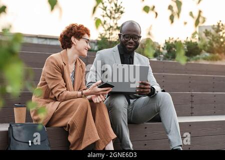 Horizontal medium long shot of successful African American man and Caucasian woman sitting together on bench in urban park looking at data on laptop Stock Photo