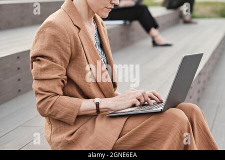 Horizontal side view shot of unrecognizable Caucasian businesswoman wearing fashionable suit sitting outdoors texting something on laptop Stock Photo