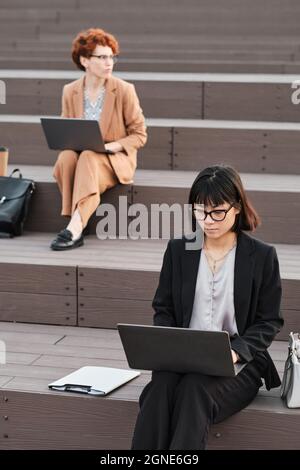 Vertical shot of two female office workers sitting on wooden stair benches outdoors on summer day using laptops Stock Photo
