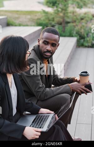 Vertical high angle shot of young African American man and unrecognizable woman sitting together outdoors drinking coffee, talking and watching someth Stock Photo