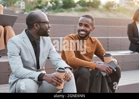 Horizontal medium shot of two successful handsome African American men wearing elegant outfits spending coffee break time relaxing outdoors chatting a Stock Photo