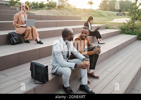 Horizontal high angle view shot of modern ethnically diverse men and women spending time outdoors in urban park working and having coffee break Stock Photo