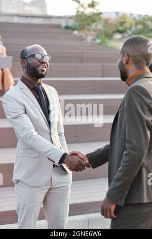 Vertical medium long shot of two successful handsome African American businessmen greeting each other with handshake outdoors Stock Photo