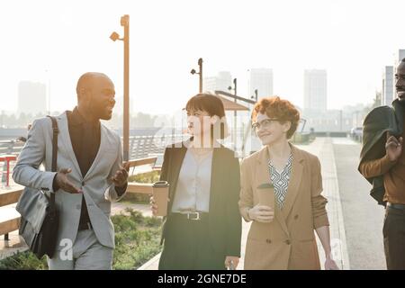 Horizontal medium long shot of four young multi-ethnic colleagues strolling down street together drinking fresh coffee and talking about various thing Stock Photo