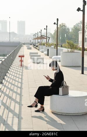 Vertical long shot of beautiful young Asian woman wearing stylish elegant outfit sitting outdoors surfing Internet on smartphone, copy space Stock Photo