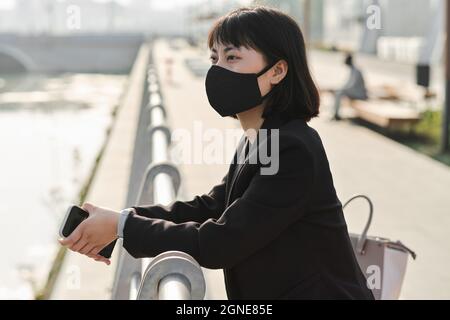Horizontal medium portrait of modern Asian woman wearing protective mask on face standing on embankment looking at river and thinking of something Stock Photo