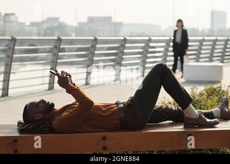 Horizontal side view shot of modern African American man relaxing on wooden bench outdoors watching something on smartphone Stock Photo