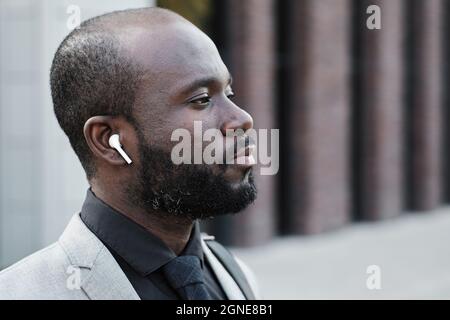Horizontal side view close-up portrait of serious African American man wearing earbuds standing outdoors listening to music thinking about something Stock Photo