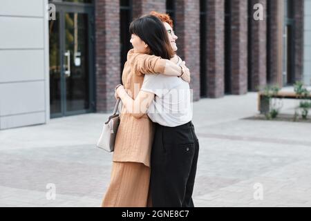 Horizontal medium long shot of modern Caucasian and young Asian women greeting each other with hug outdoors Stock Photo