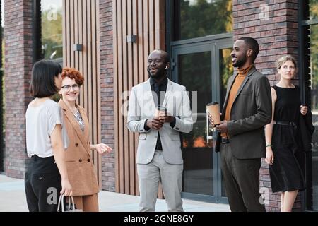 Medium long shot of multi-ethnic co-workers standing outdoors drinking coffee and chatting about funny things during break Stock Photo