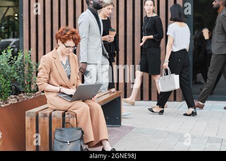 Portrait of serious mature businesswoman sitting on wooden bench outdoors in business district planning her work using laptop Stock Photo