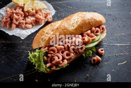 From above of appetizing submarine sandwich with crispy baguette bread stuffed with shrimps and fresh cucumber slices with lettuce on black surface Stock Photo