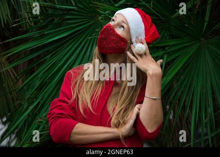 Caucasian blond girl in red hat, coat and red protective face mask with palm leafs behind. Thinking of Christmas 2020. Celebrating pandemic new 2021 Stock Photo