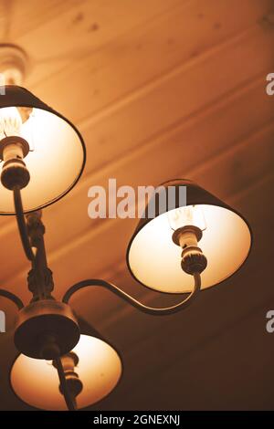 Beautiful elegant retro-style lamps hang on the background of the wooden ceiling and exude a warm cozy light at night. Hotel. Stock Photo