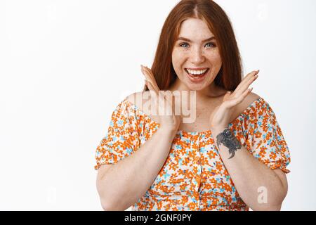 Excited redhead girl claps hands and smiles, looking amazed at camera, standing over white background Stock Photo