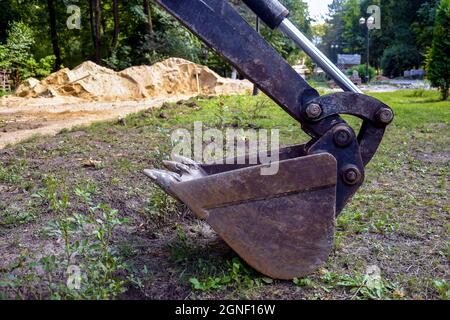 Bucket of small excavator is located on lawn. Strong teeth are ready to bite into the ground. Construction work in the park. Close-up. Selective focus Stock Photo