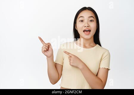 Enthusiastic korean girl pointing fingers left, looking amazed at camera, standing in yellow t-shirt over white background Stock Photo