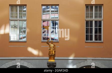 a beige building with three windows reflecting neighboring buildings. In front of them is a gilded statue of a knight. Riga, Latvia Stock Photo