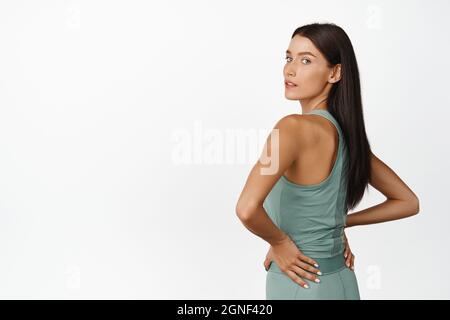 Rear view of strong and fit fitness woman turn back at camera, showing muscles, standing in workout clothes over white background Stock Photo