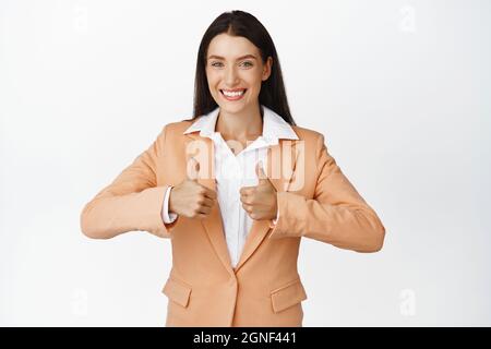Smiling businesswoman showing thumbs up, approve smth good. Saleswoman give positive feedback, standing in suit against white background Stock Photo