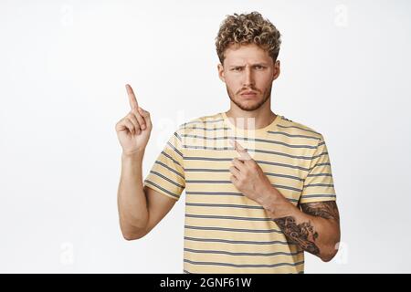 Disappointed blond man showing bad advertisement, pointing at upper left corner, frowning and expressing dislike with angry face expression, white Stock Photo