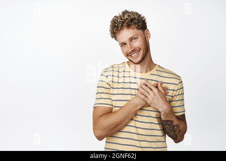 Cute blond guy smiles thankful, holds hands on chest and looks happy, has something in his heart, sweet memories gesture, standing over white Stock Photo