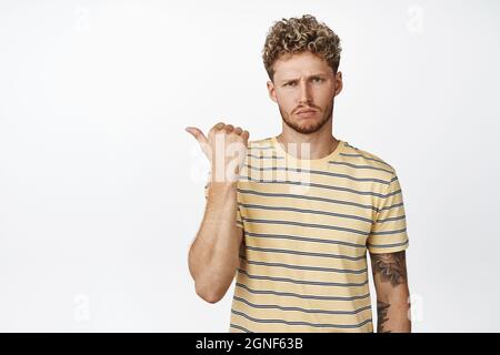 Upset young blond guy pointing finger left, looking disappointed and frowning at camera, white background Stock Photo