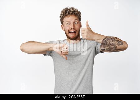 Indecisive funny guy making crazy eyes, showing tongue, thumbs up and thumbs down, cant make decision, rate something, give feedback, like or dislike Stock Photo