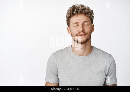 Close up portrait of dreamy blond man close eyes, thinking of something, daydreaming, imaging in his mind, standing in gray t-shirt over white Stock Photo