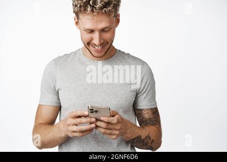 Portrait of handsome blond caucasian man using mobile phone and smiling, looking at his smartphone screen, chatting on cellphone, standing in gray t Stock Photo