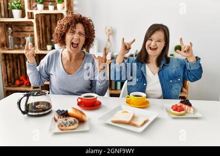 Family of mother and down syndrome daughter sitting at home eating breakfast shouting with crazy expression doing rock symbol with hands up. music sta Stock Photo