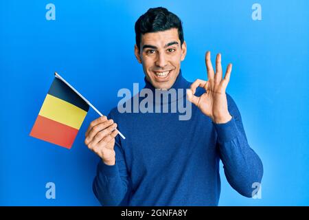 Handsome hispanic man holding belgium flag doing ok sign with fingers, smiling friendly gesturing excellent symbol Stock Photo