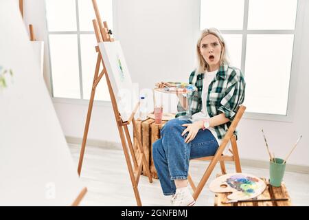 Young artist woman painting on canvas at art studio in shock face, looking skeptical and sarcastic, surprised with open mouth Stock Photo