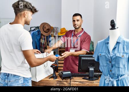 Young hispanic customer man paying to shopkeeper using credit card at clothing store. Stock Photo