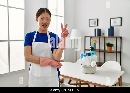 Young chinese girl wearing cleaner uniform standing at home smiling with happy face winking at the camera doing victory sign. number two. Stock Photo