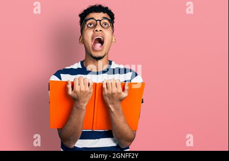 Young african american man reading a book with glasses angry and mad screaming frustrated and furious, shouting with anger looking up. Stock Photo
