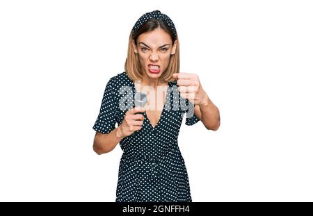 Young blonde girl singing song using microphone annoyed and frustrated shouting with anger, yelling crazy with anger and hand raised Stock Photo