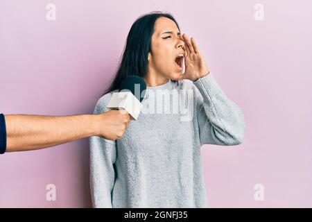 Young hispanic girl being interviewed by reporter holding microphone shouting and screaming loud to side with hand on mouth. communication concept. Stock Photo