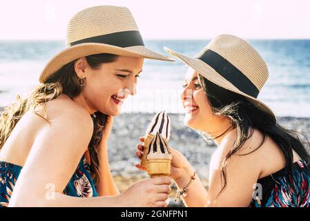 Two beautiful female friends in straw hat having fun enjoying cold ice cream cones on a peaceful beach during summer holiday. Cheerful women enjoying Stock Photo