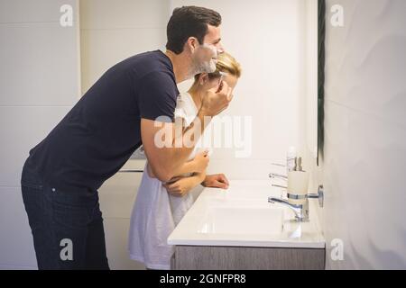 Joyful couple having fun in the bathroom during morning while man is shaving his beard using razor and foam. Happy couple getting ready together in th Stock Photo