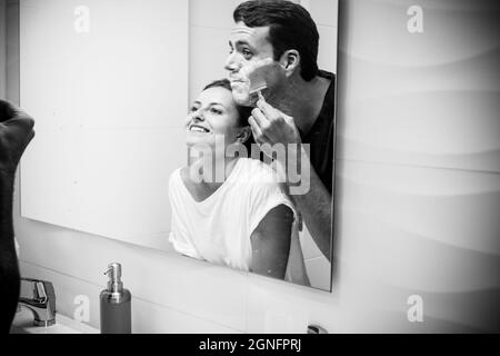 Joyful couple having fun in the bathroom during morning while man is shaving his beard using razor and foam. Happy couple getting ready together in th Stock Photo
