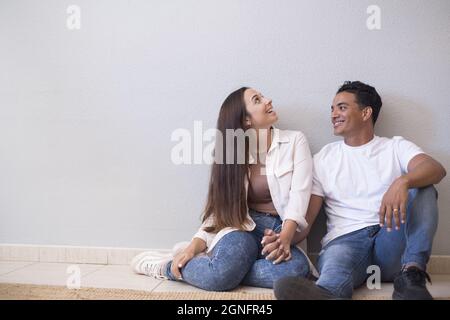 Young couple sitting on floor of living room holding hands and leaning on wall. Couple spending quality time at home. Cheerful couple sitting on floor Stock Photo
