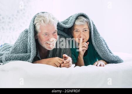 Senior couple lying on front and gesturing finger on lips on bed. Elderly couple spending leisure with each other. Happy old couple lying on cozy bed Stock Photo