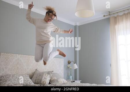 Crazy young woman jumping on bed at home. Overjoyed woman going crazy. Carefree woman dancing and jumping on bed in excitement celebrating success at Stock Photo